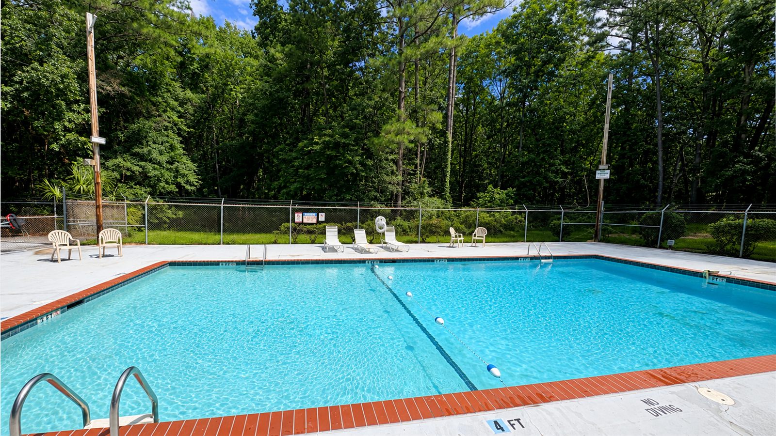 The Winding Creek community pool on a sunny day, clear blue water bordered by red brick coping, poolside chairs and tall pines behind the fence.